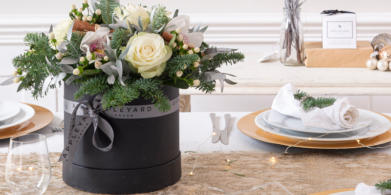 Christmas hatbox with white roses and orchids on a table next to a plate and glass. 