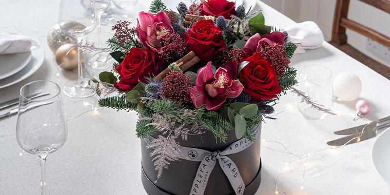 A floral hatbox with orchids, roses, cinnamon, pine & eryngium on a table with glasses and cutlery.