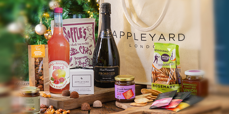 A hamper of food sits in front of a christmas tree, an Appleyard bag is behind the food and alcohol