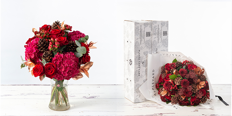 A red toned bouquet to the left in a vase, containing prominent hydrangeas and roses.To the left of this a hand tied bouquet of similar colours lies in its wrapping next to a delivery box stood on its end. This second bouquet contains red roses chryanthemums and pine cones