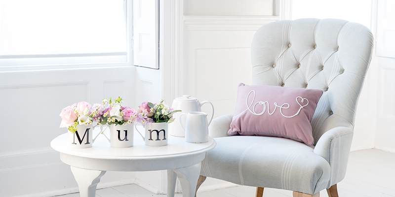 A homely scene of a comfy chair and table, the furniture is white and there is a pink cushion with the word 'love' on it sitting on the chair. There are three mugs on the table monogrammed 'M' 'u''m' and these contain pale flowers in oink white and purple