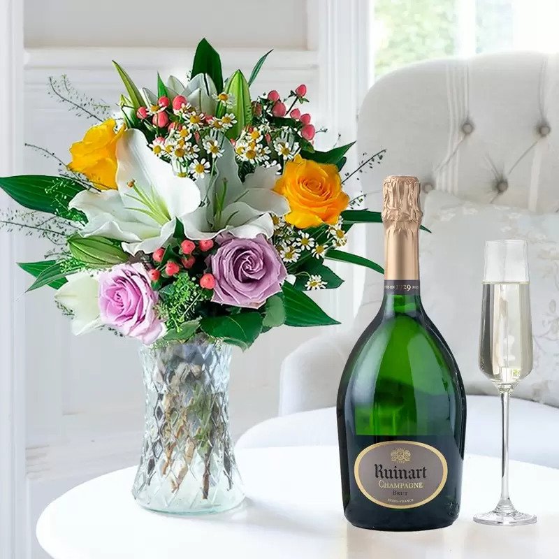 A bouquet of yellow and lilac roses and white lilies next to a bottle of Ruinart champagne with a glass.
